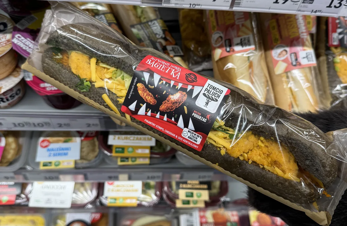 Bagietka Halloween Tomcio Paluch z czarną bułką trzymana przy lodówce z kanapkami w Żabce