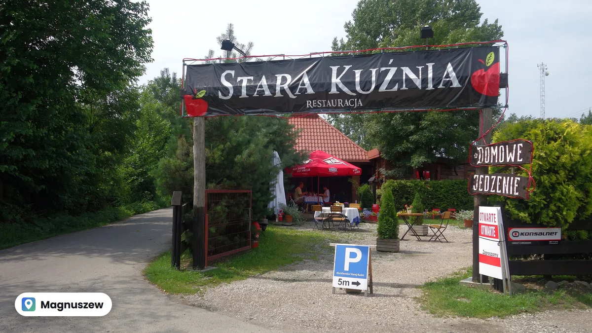 Wejście do restauracji Stara Kuźnia w Magnuszewie, nad bramą baner z nazwą, w tle ogródek ze stolikami pod parasolami