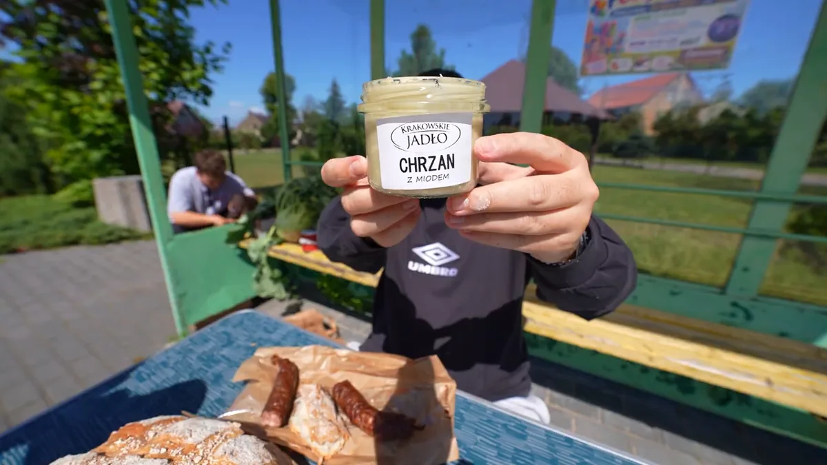Książulo przy stole przed sklepem Karolinka trzyma słoik chrzanu z miodem Krakowskie Jadło