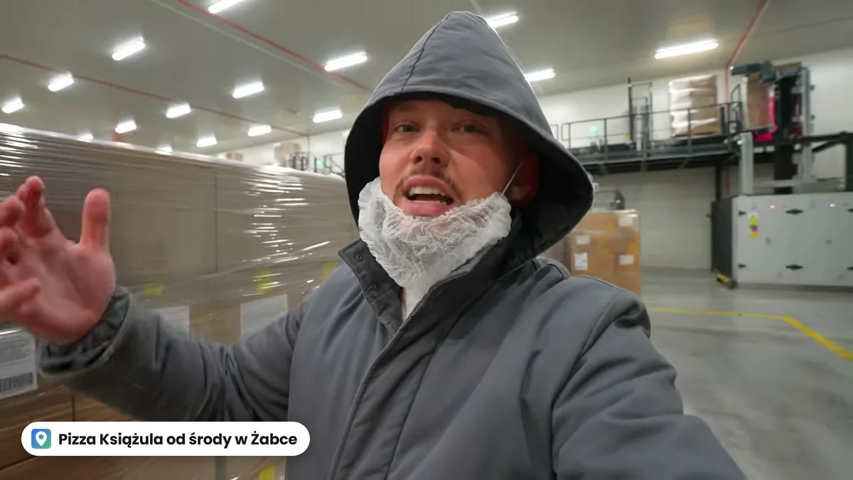 Książulo w kapturze i siateczce na brodzie w hali fabryki na Słowacji, za nim palety z kartonami pizzy