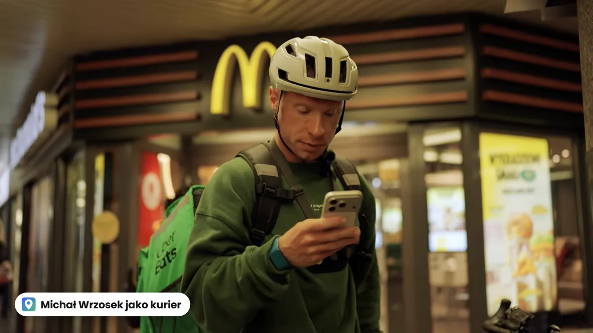 Michał Wrzosek w kasku i z torbą Uber Eats patrzy w telefon przed lokalem McDonald's, kadr z filmu