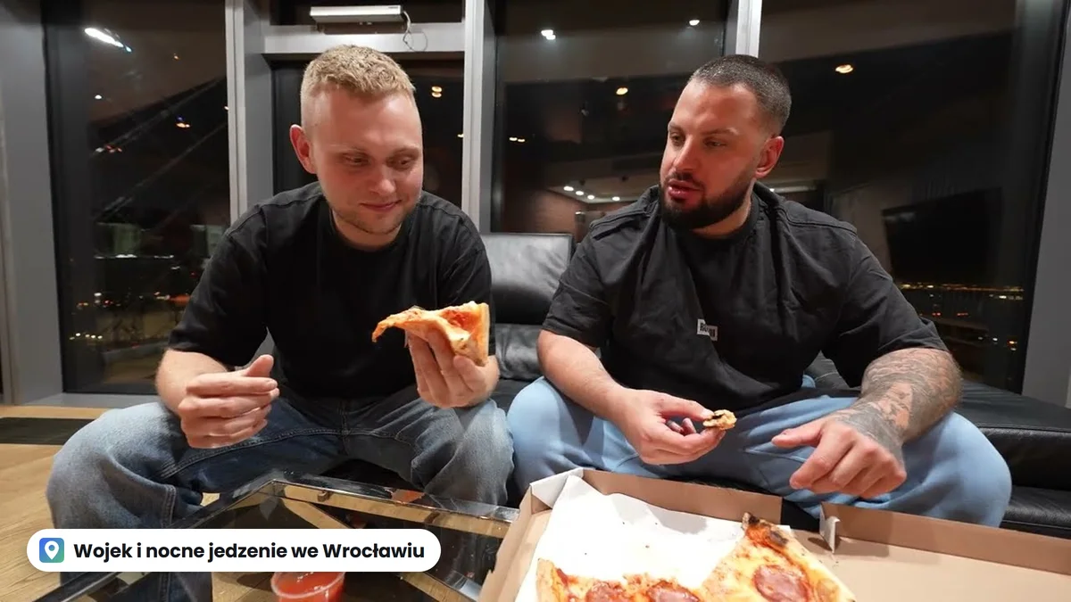Wojek i Staszek na kanapie w apartamencie w Sky Towerze nocą jedzą pizzę z pudełka, za oknem światła Wrocławia