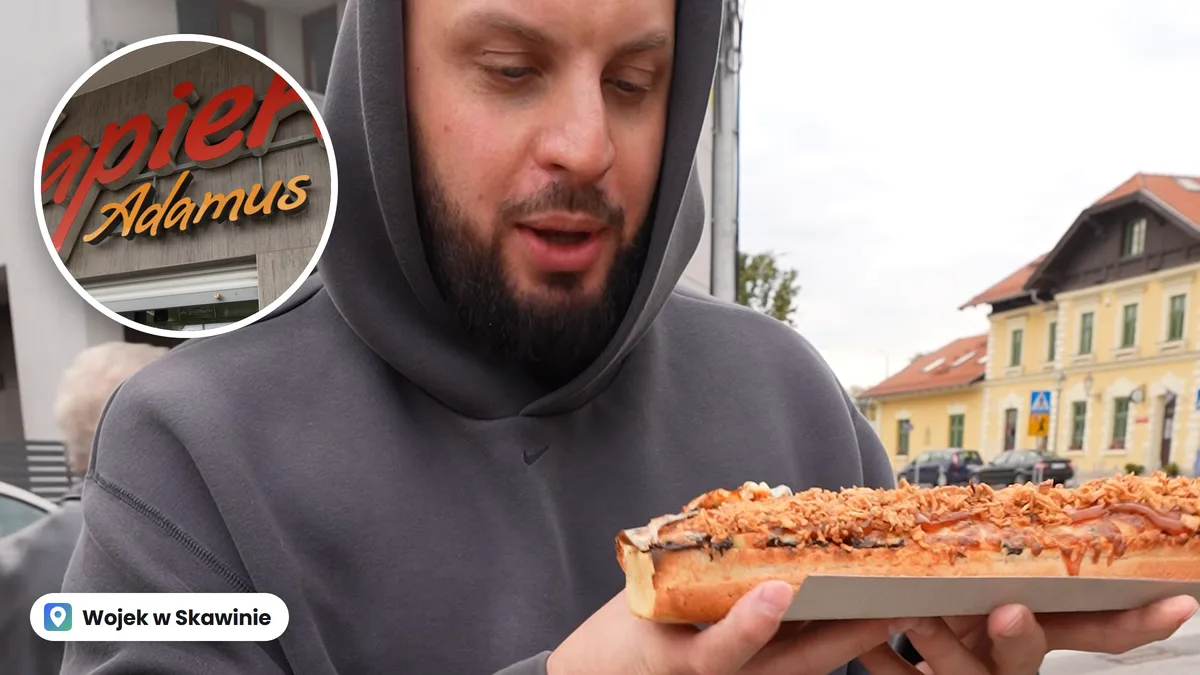 Wojek w szarej bluzie z kapturem trzyma długą zapiekankę z prażoną cebulką i keczupem, w kółku szyld Zapiekanki Adamus